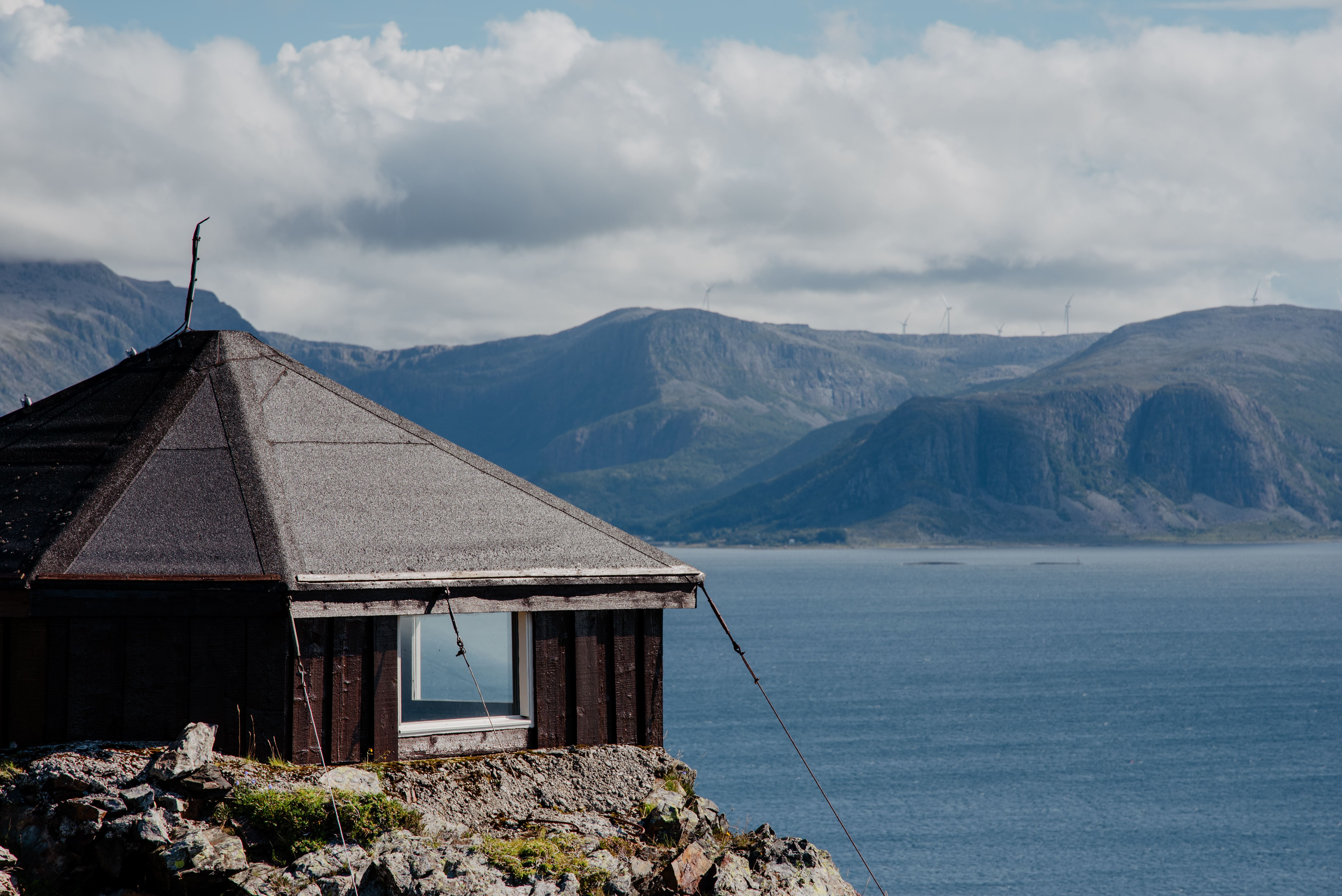 Small dark cabin with a sloped roof sits on a rocky shore, overlooking a wide fjord and distant mountains under clouds.