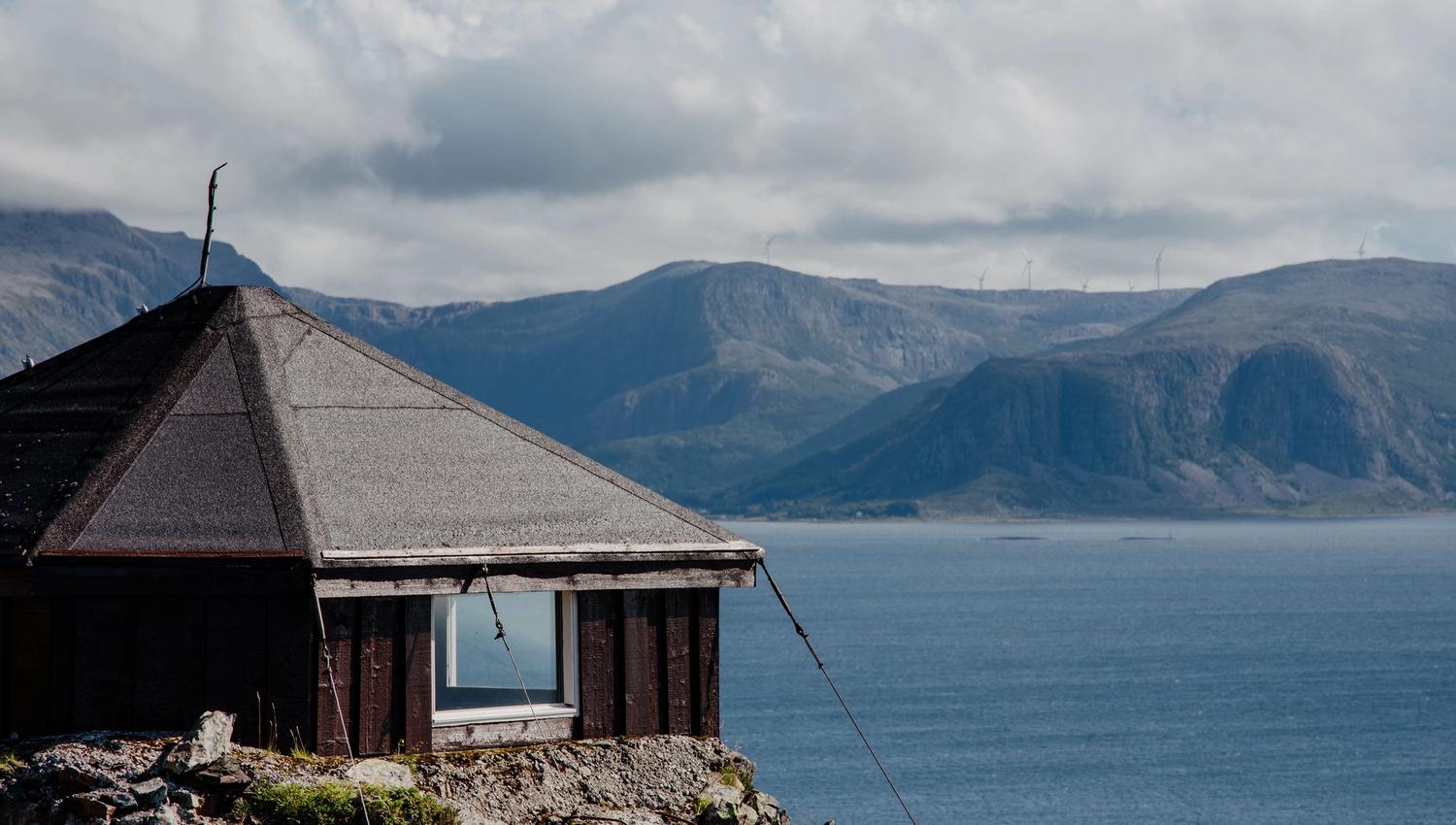 Small dark cabin with a sloped roof sits on a rocky shore, overlooking a wide fjord and distant mountains under clouds.