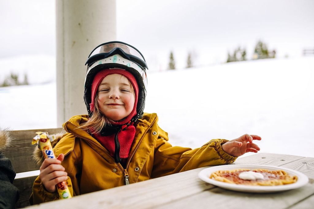 Person in a helmet and goggles sits at an outdoor table in snowy surroundings, holding a snack beside a plate.