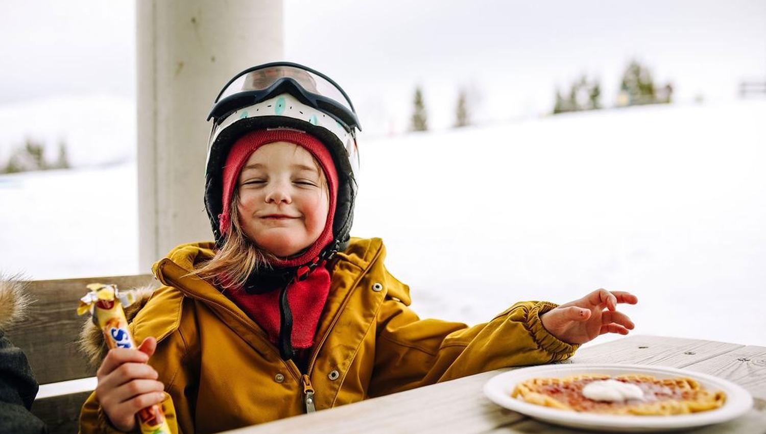 Person in a helmet and goggles sits at an outdoor table in snowy surroundings, holding a snack beside a plate.
