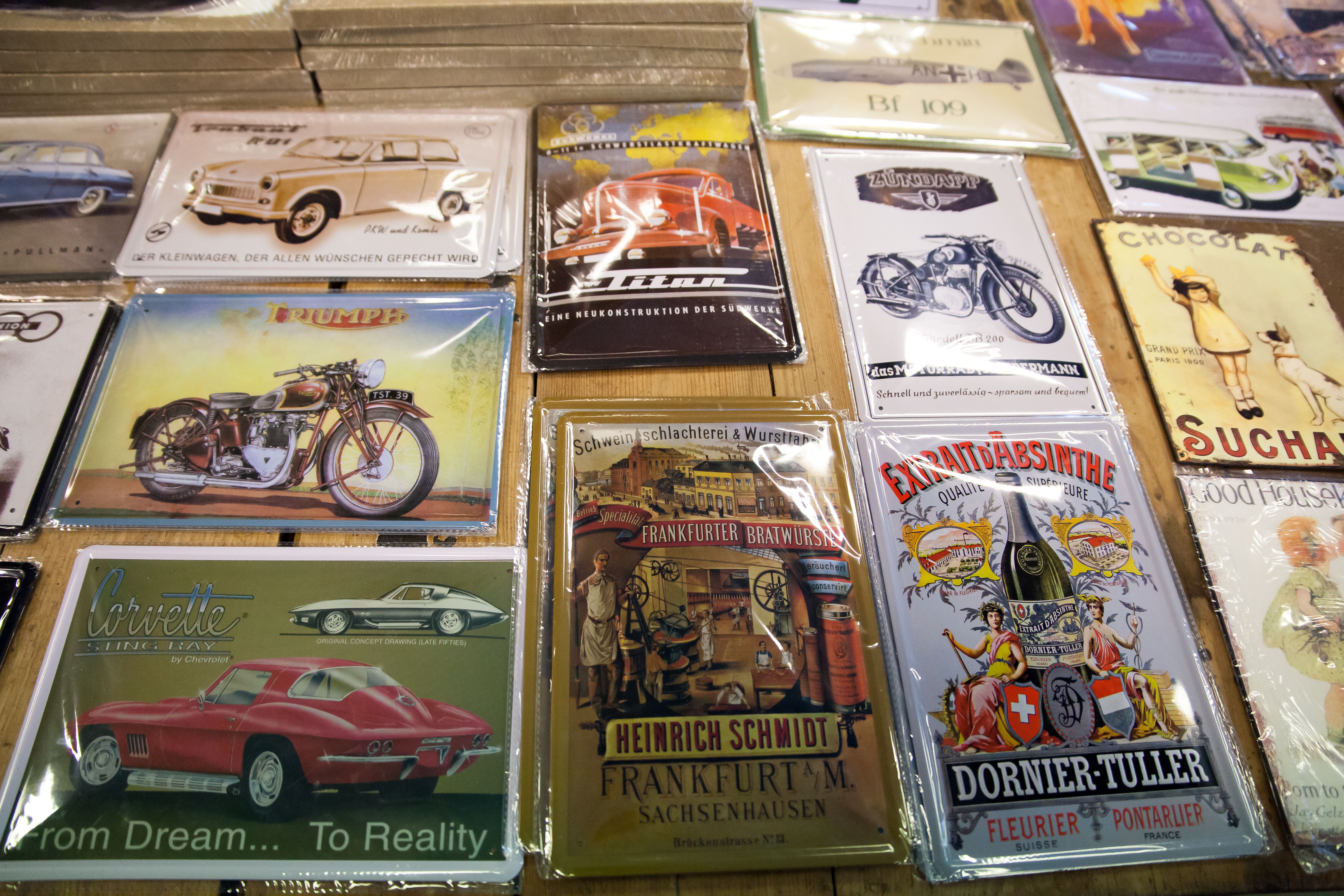 Metal signs for sale. The images feature vintage cars and motorcycles, drinks, and more. Among the motifs are:

A reddish-brown Triumph

A red Corvett