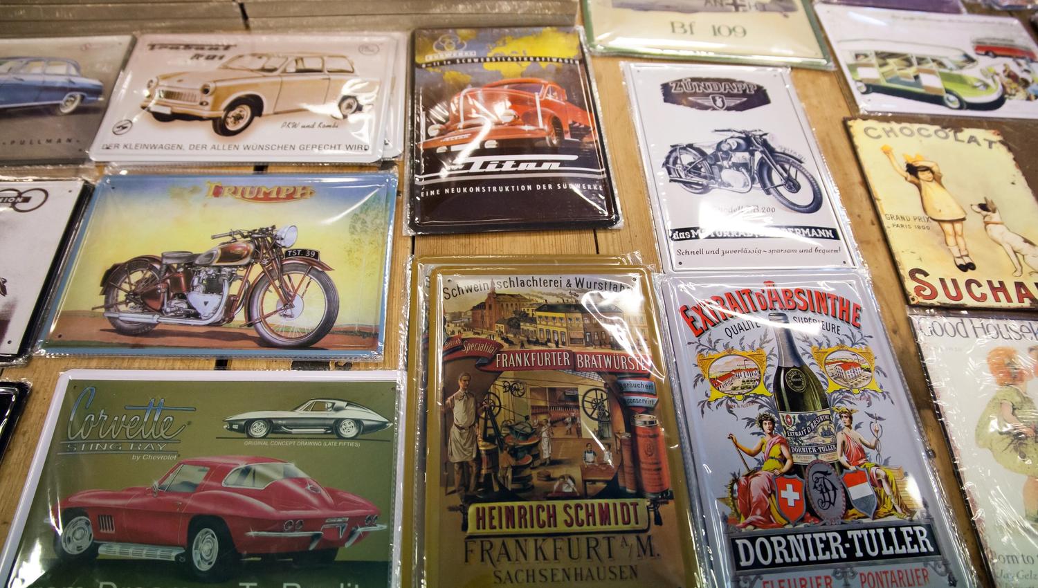 Metal signs for sale. The images feature vintage cars and motorcycles, drinks, and more. Among the motifs are:
A reddish-brown Triumph
A red Corvett