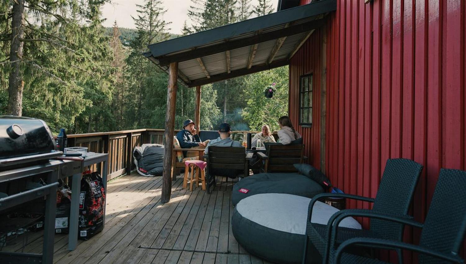 Terrasse ved en rød trebygning i skogsomgivelser, med sittegruppe og grill klart til bruk.