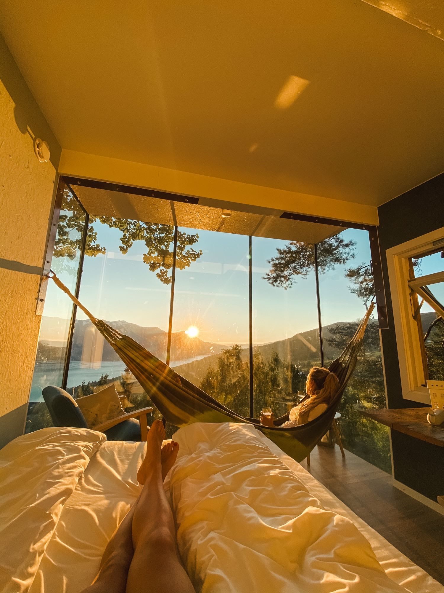 Cozy room with bed and two hammocks facing a glass wall, overlooking a lake, trees, and mountains at sunset..
