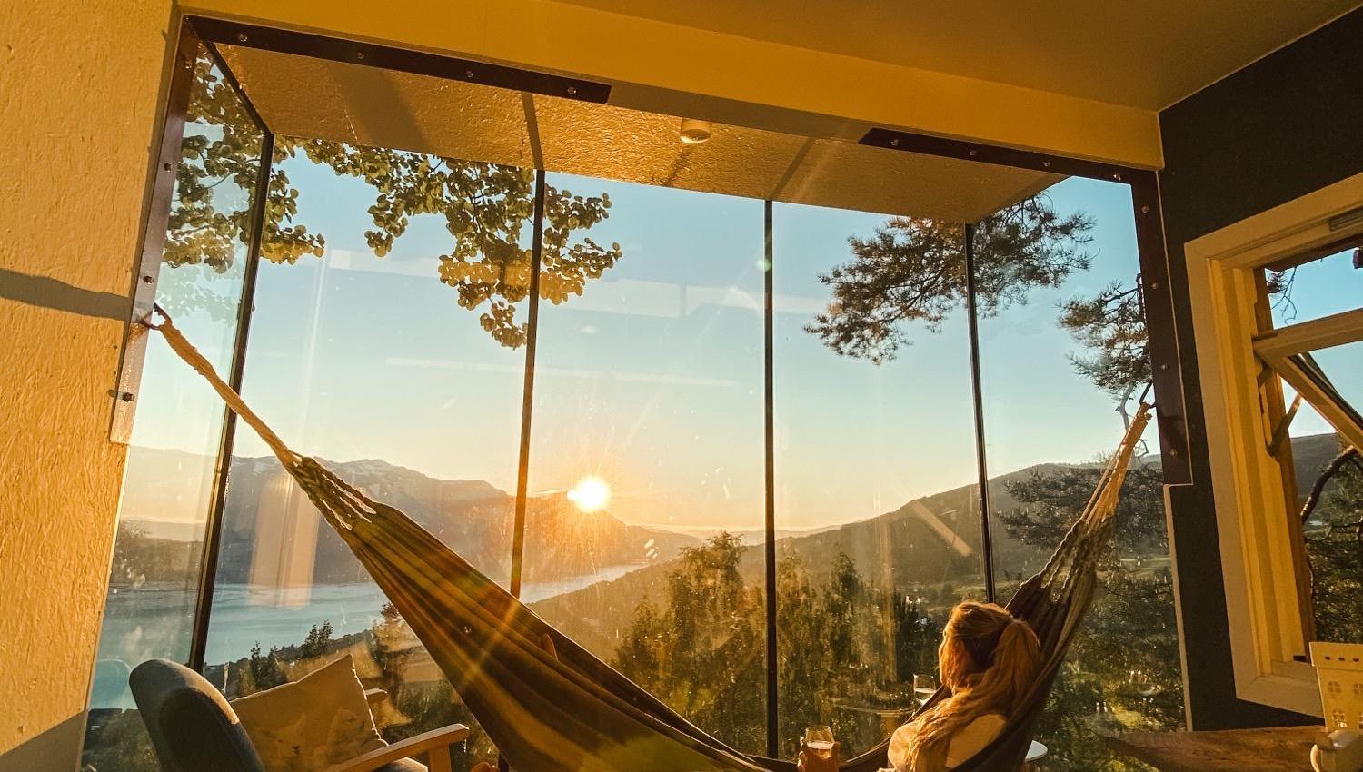 Cozy room with bed and two hammocks facing a glass wall, overlooking a lake, trees, and mountains at sunset..