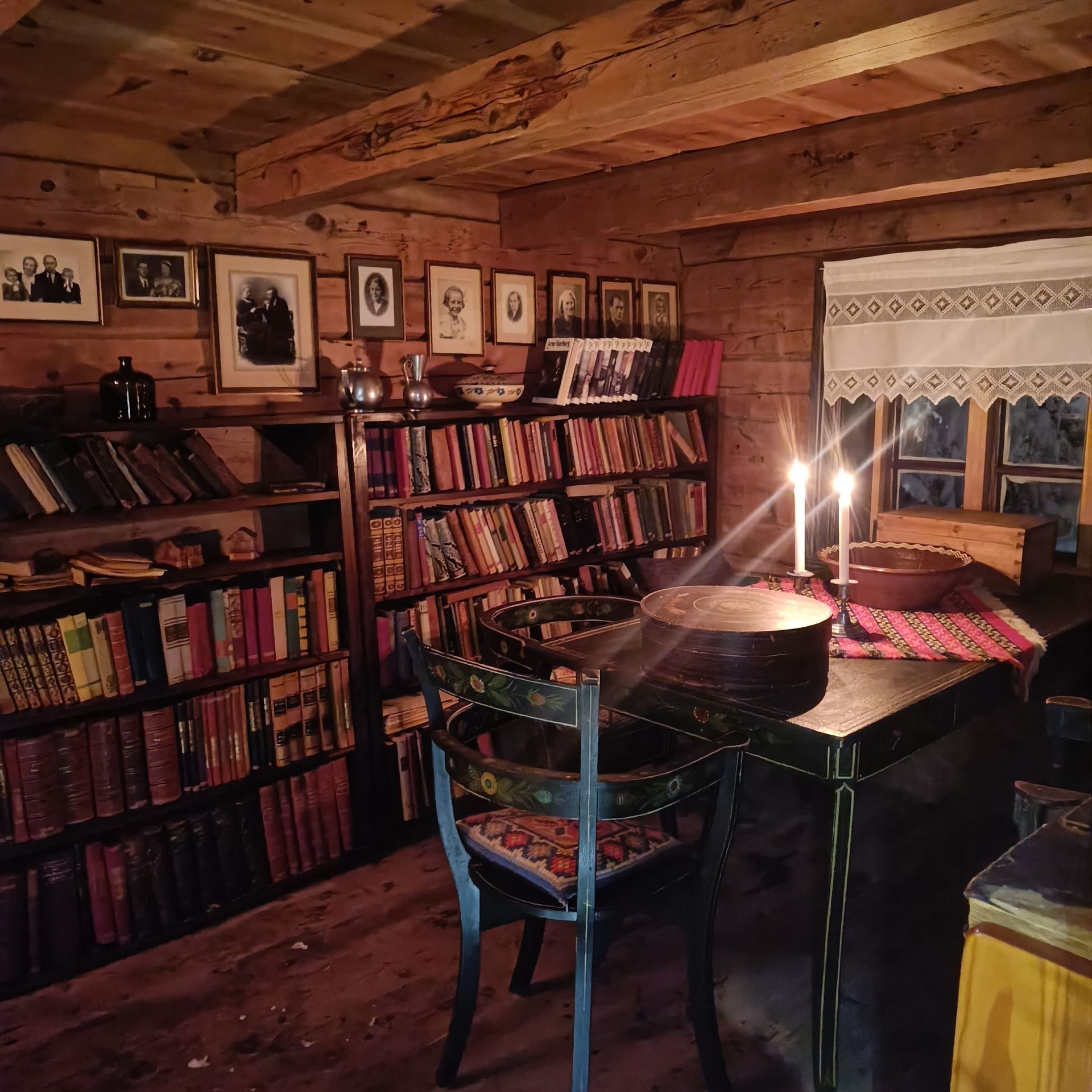 Orheimstunet in Nordfjord - Rustic wooden room with bookshelves, framed portraits, a table with two lit candles, and a window with a lace curtain..