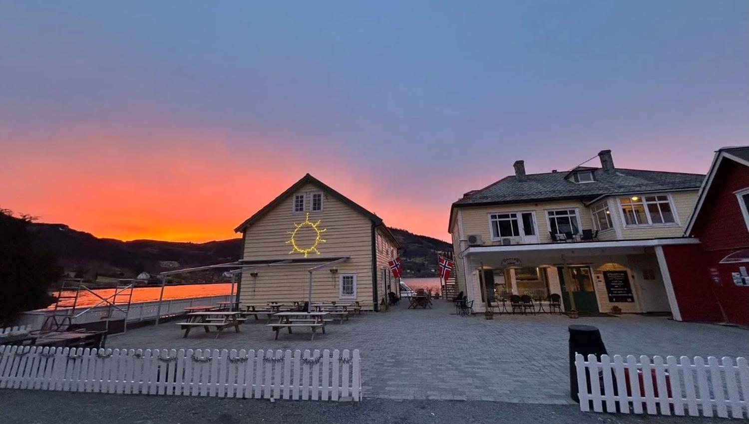 Two wooden buildings by a waterfront with picnic tables and a white fence under a vivid orange sunset sky at Davik Fjordparadis in Bremanger..