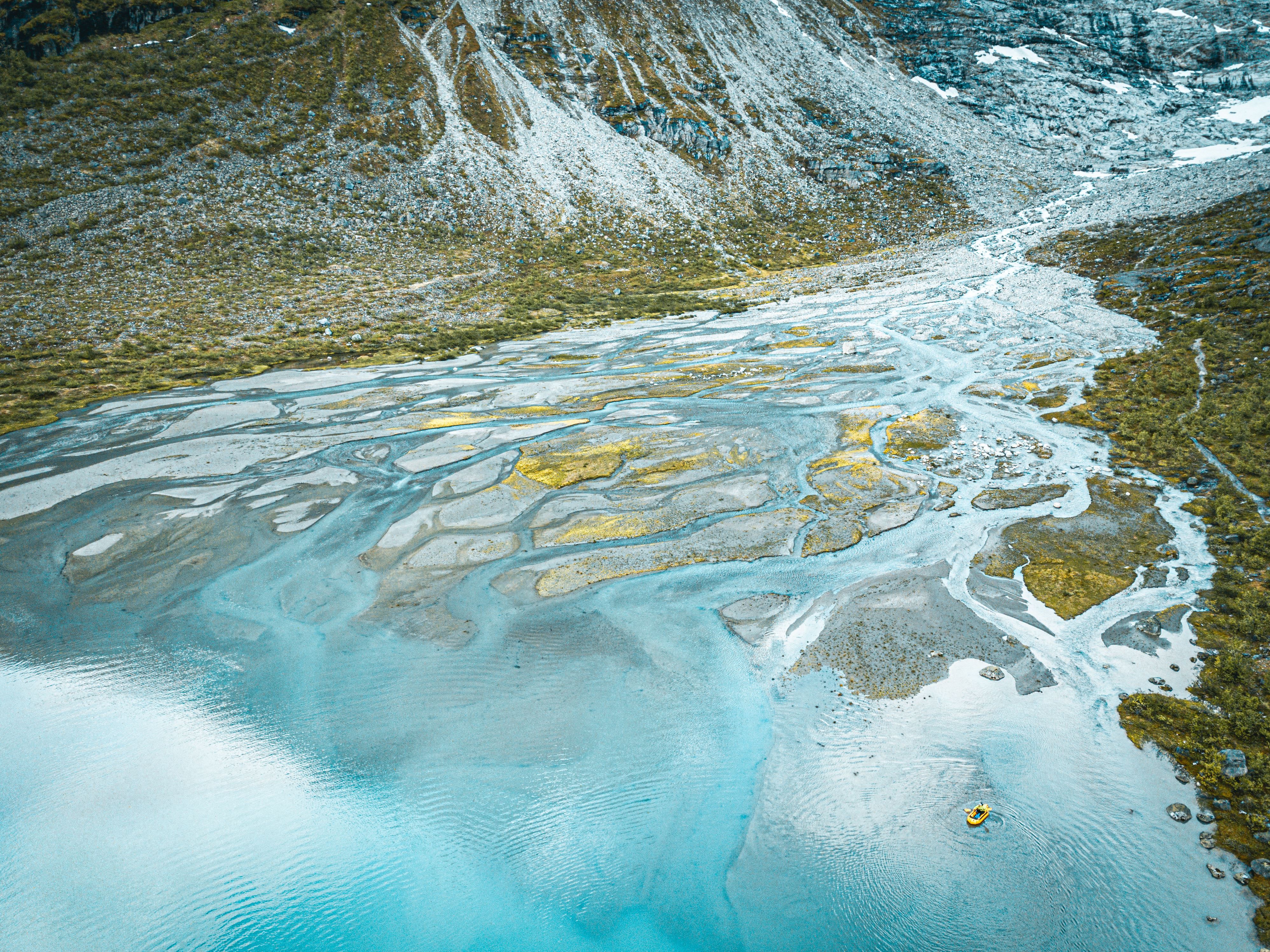 Aerial view of turquoise braided river channels and gravel bars flowing into a lake, with mountains behind.
