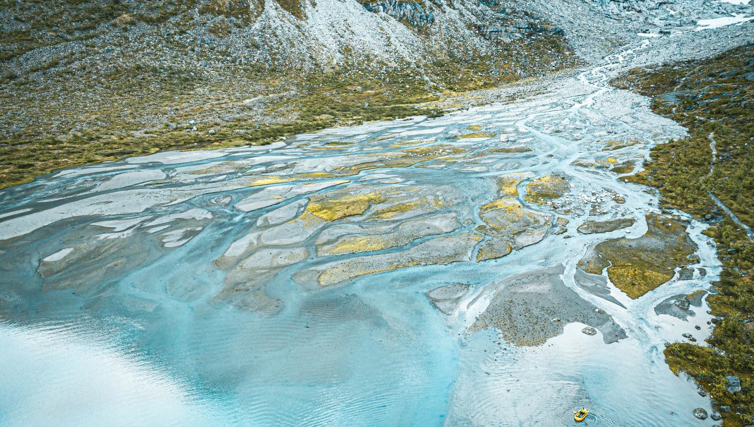Aerial view of turquoise braided river channels and gravel bars flowing into a lake, with mountains behind.