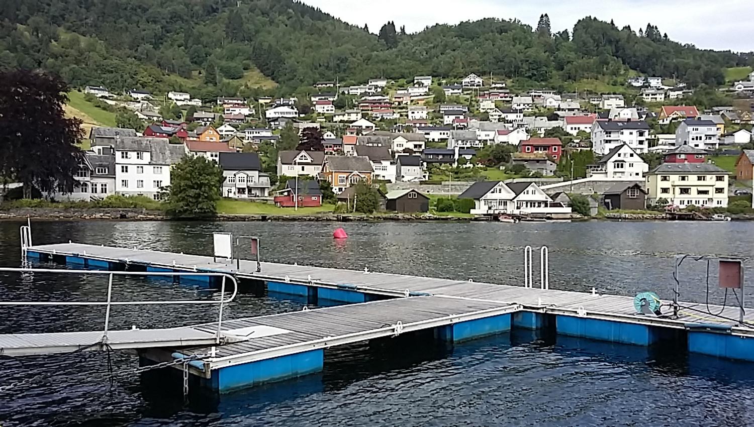 View from the dock at Norheimsund Guest Harbour towards white houses and green hills surrounding the fjord.