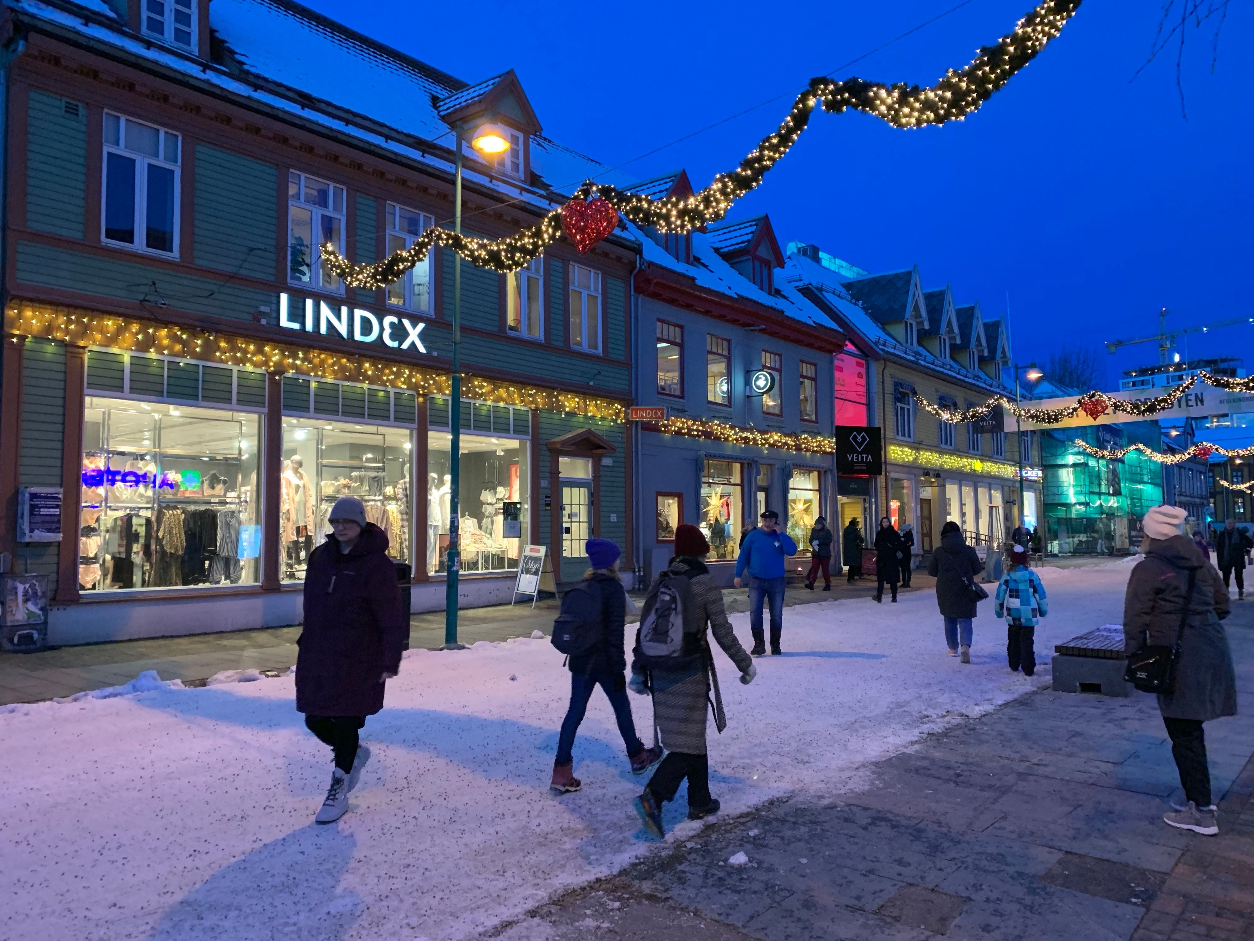 Folk går i snødekt gate med julelys og butikker en vinterkveld