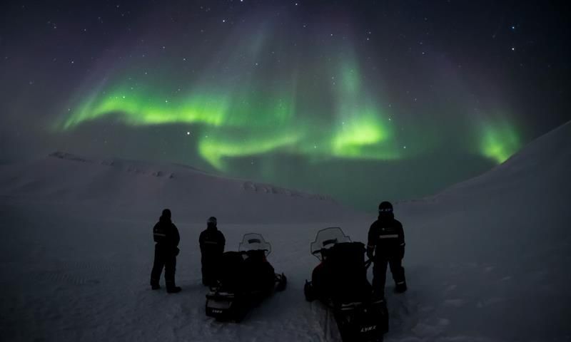 Tre personer står ved siden av to snøscootere og ser på nordlyset