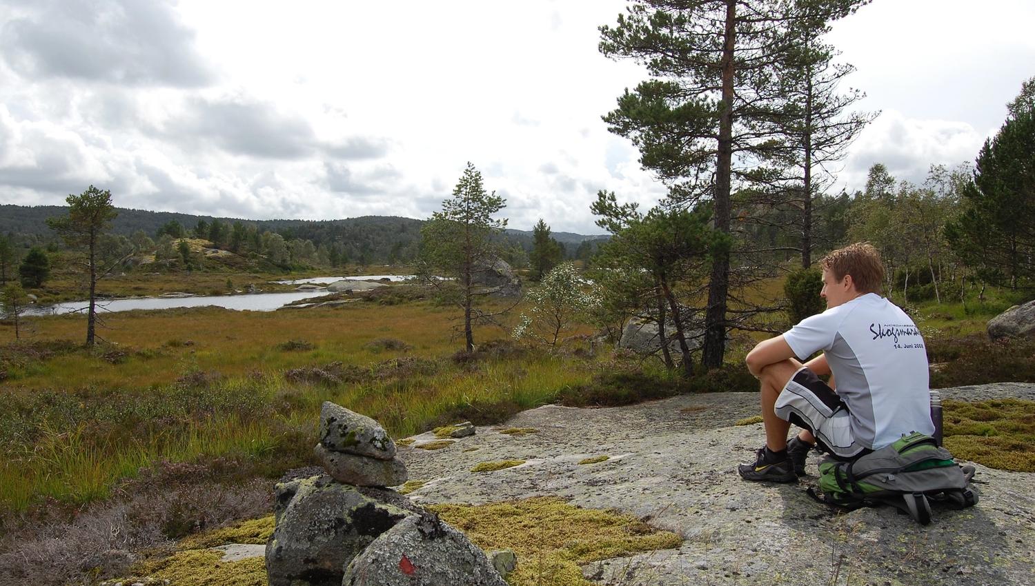A man sitting on his backpack, looking out over the landscape with a water pond.