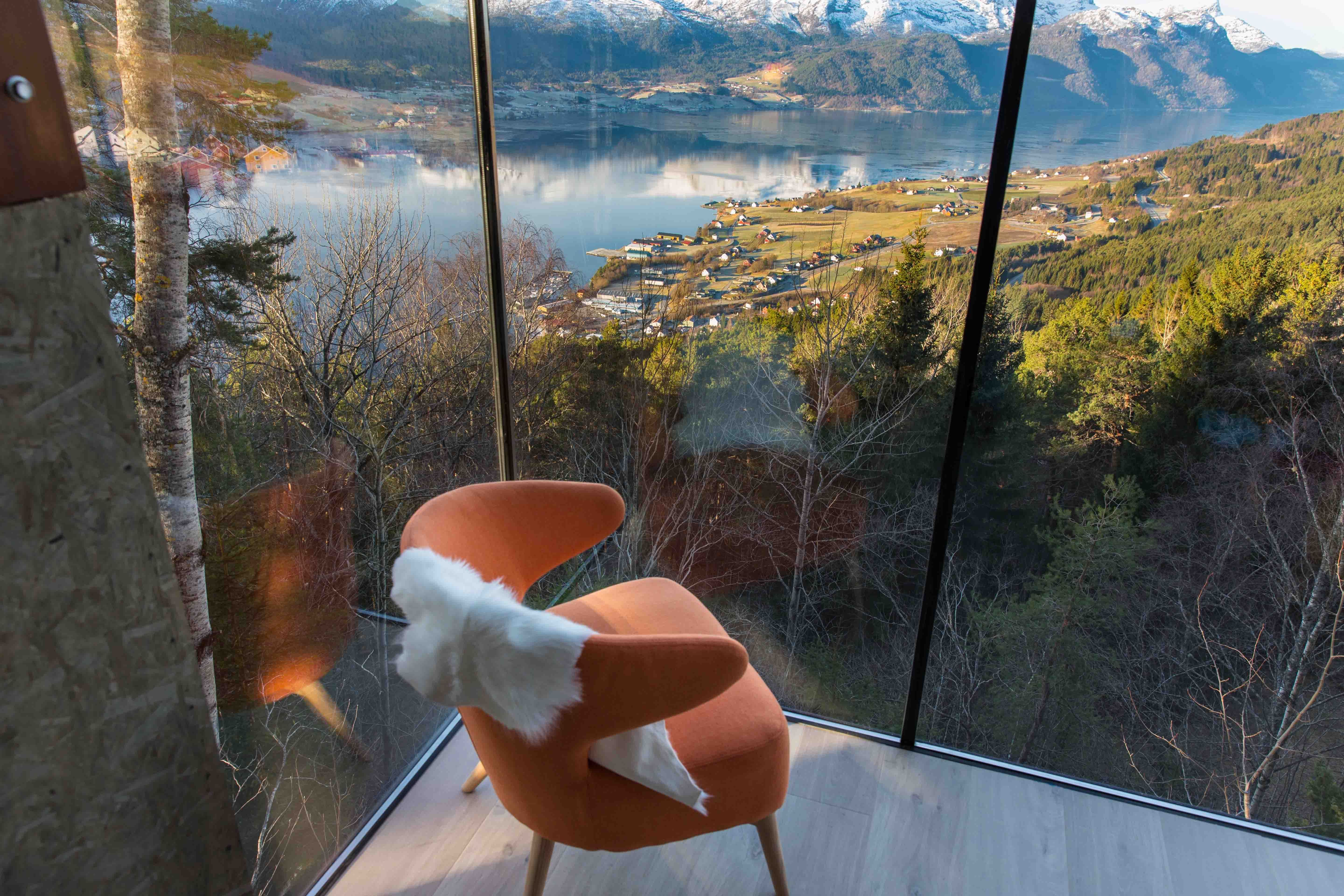 Orange armchair with white throw beside a glass wall, overlooking a fjord, village fields, forest, and snowy mountains..