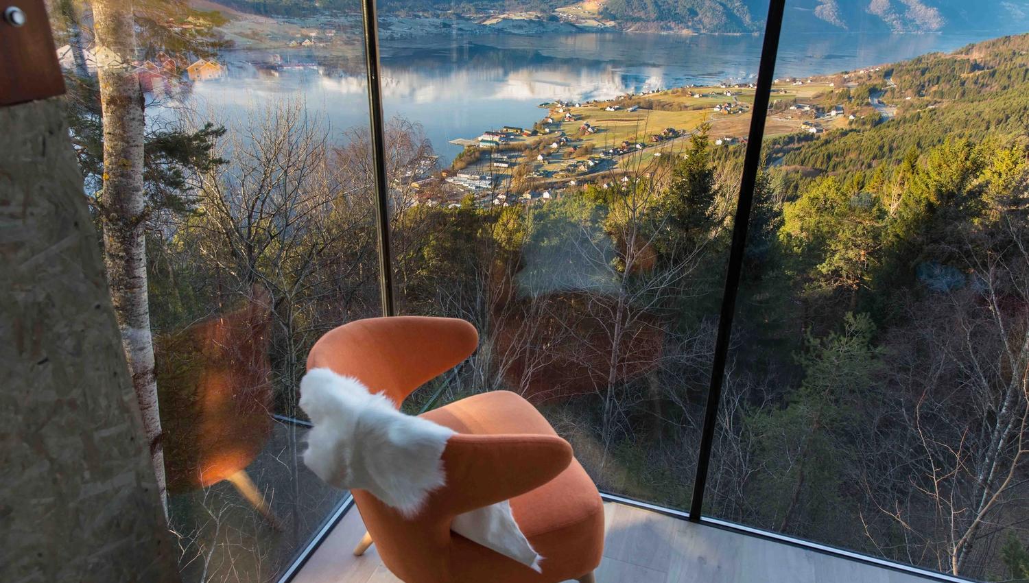 Orange armchair with white throw beside a glass wall, overlooking a fjord, village fields, forest, and snowy mountains..