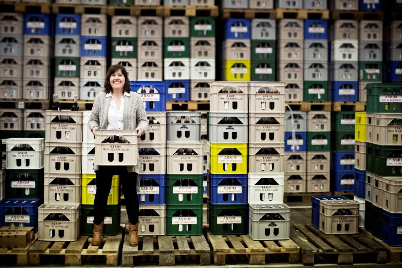 A woman stands in a warehouse filled with crates stacked in tall rows. She is smiling and holding a crate with bottles in front of her. The crates are