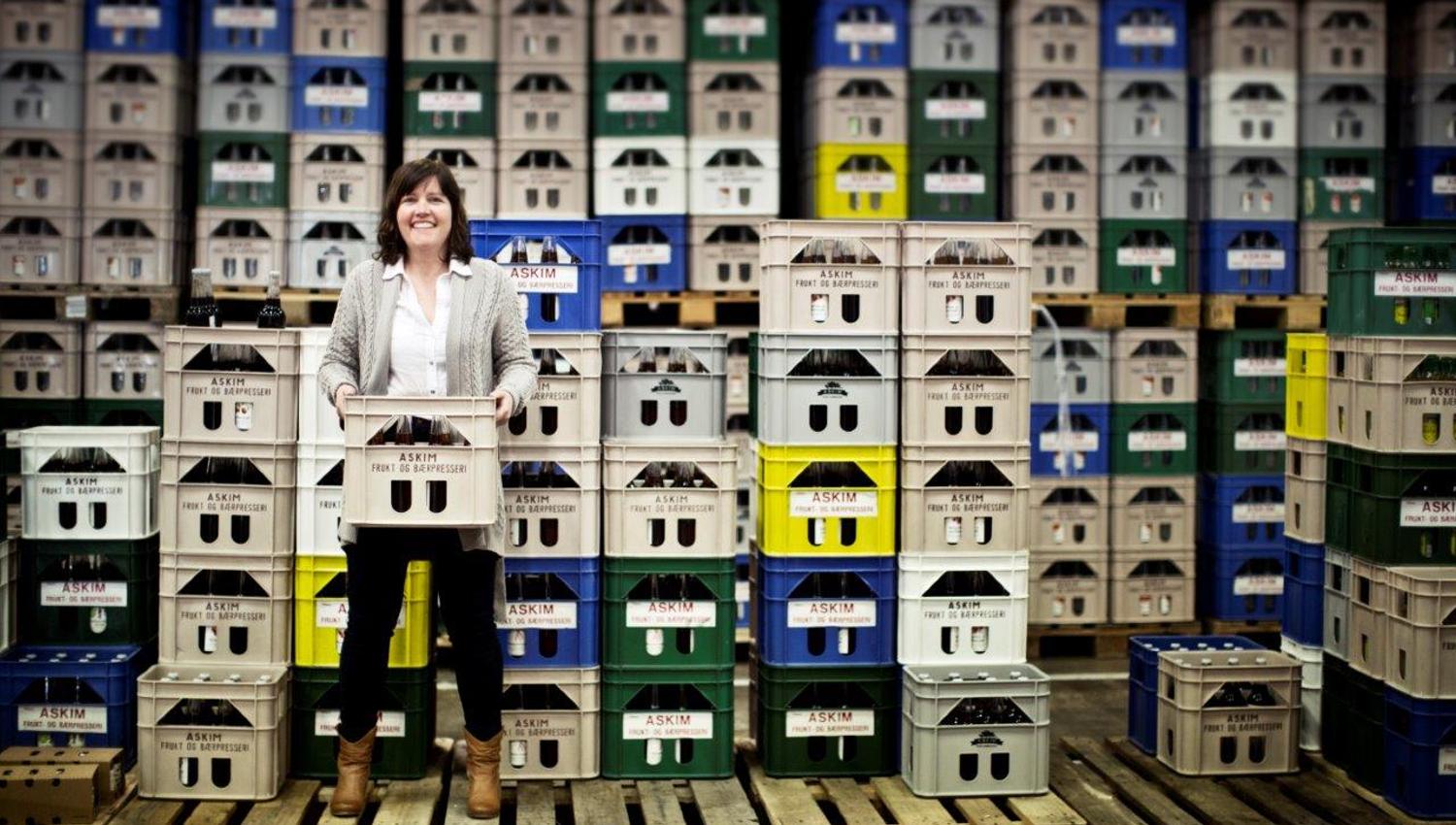 A woman stands in a warehouse filled with crates stacked in tall rows. She is smiling and holding a crate with bottles in front of her. The crates are