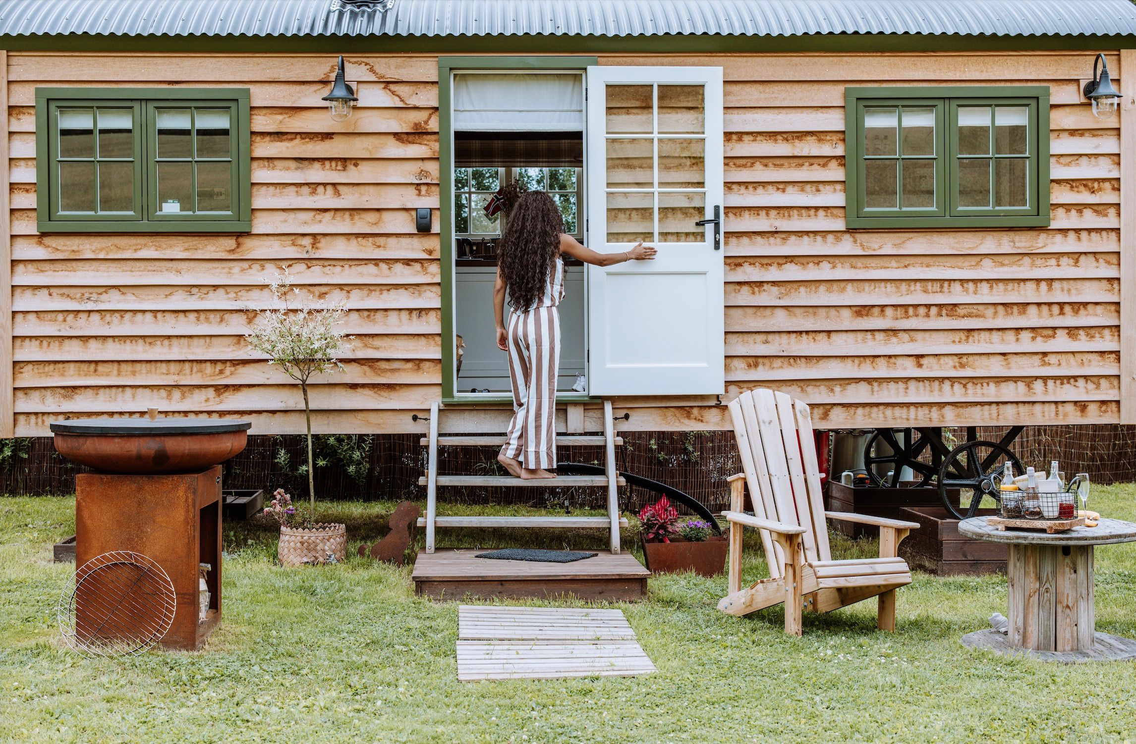girl walking into a Small wooden house with green windows and chairs in front 
