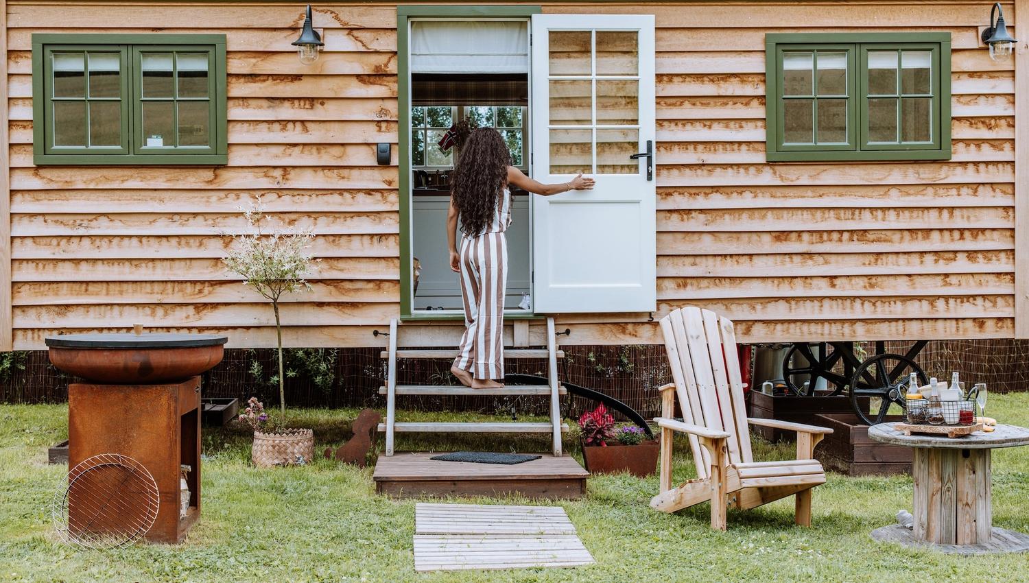 girl walking into a Small wooden house with green windows and chairs in front