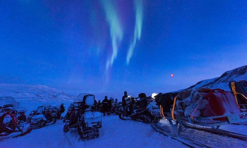 People with snowmobiles are taking a breake to watch the northern lights on the sky
