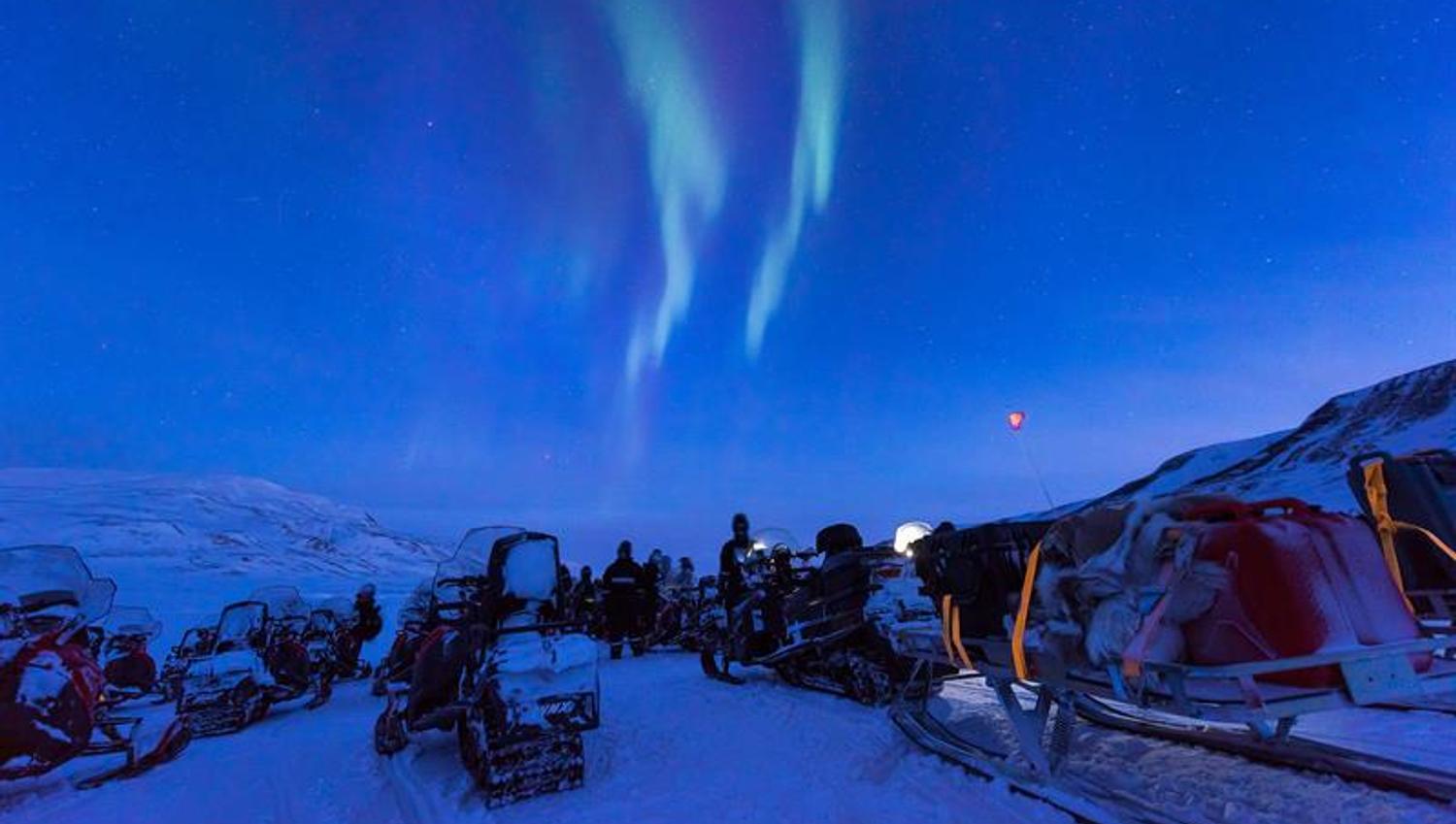 People with snowmobiles are taking a breake to watch the northern lights on the sky