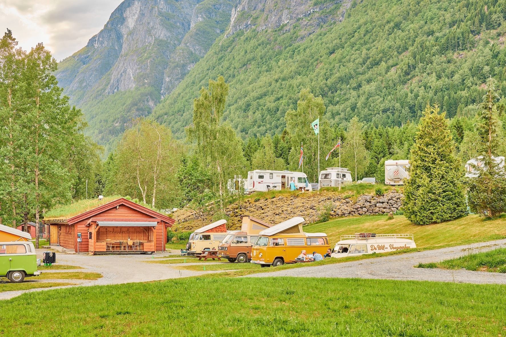 Campingplass med raud hytte, veteranbussar og bubilar i grønt landskap, med skogkledde fjell i bakgrunnen..