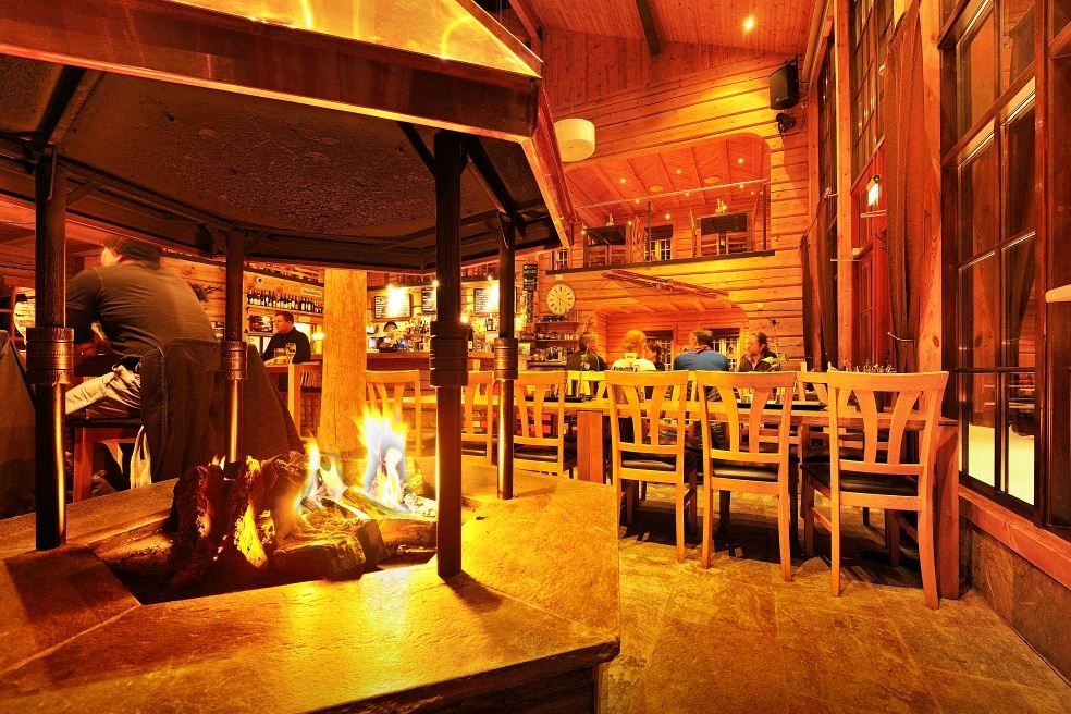 Rustikk restaurant i tre med ildsted i forgrunnen, bord og stoler, bar i bakgrunnen og gjester som sitter..