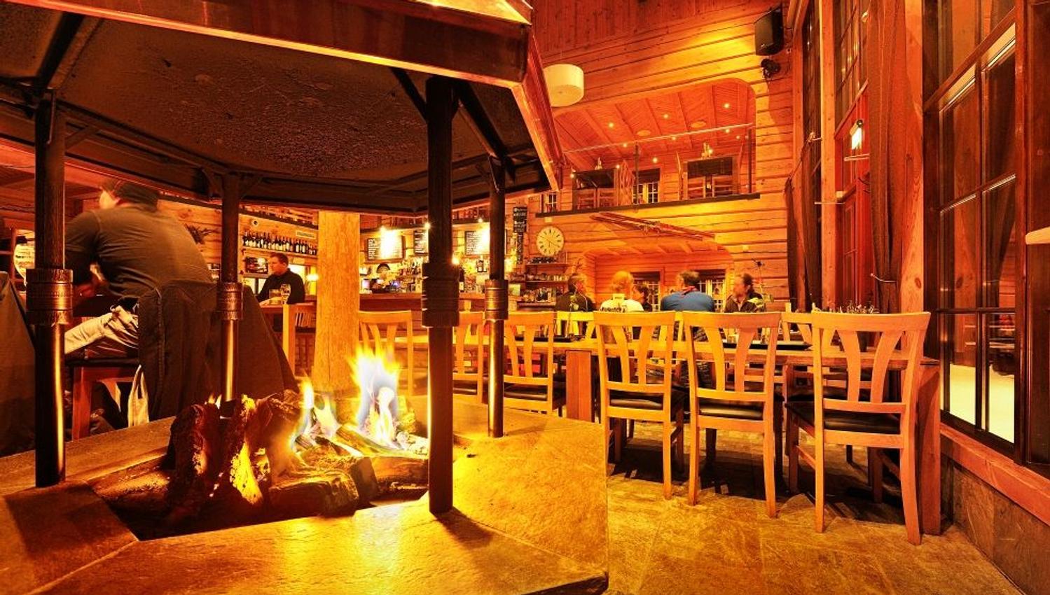 Rustikk restaurant i tre med ildsted i forgrunnen, bord og stoler, bar i bakgrunnen og gjester som sitter..