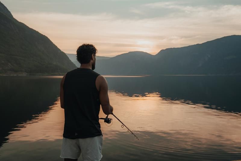 A man is fishing by the fjord during sunset.