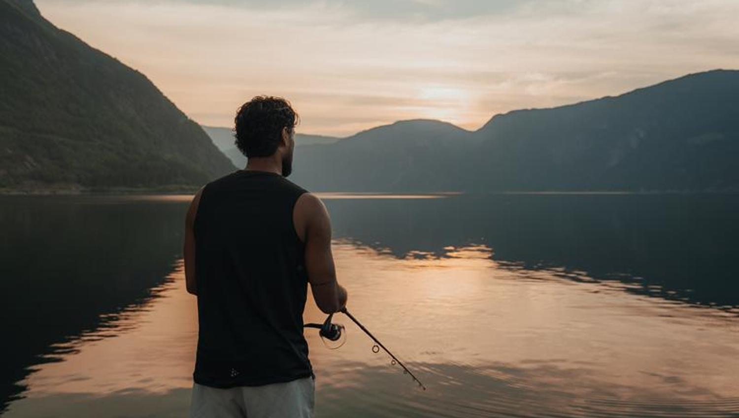 A man is fishing by the fjord during sunset.
