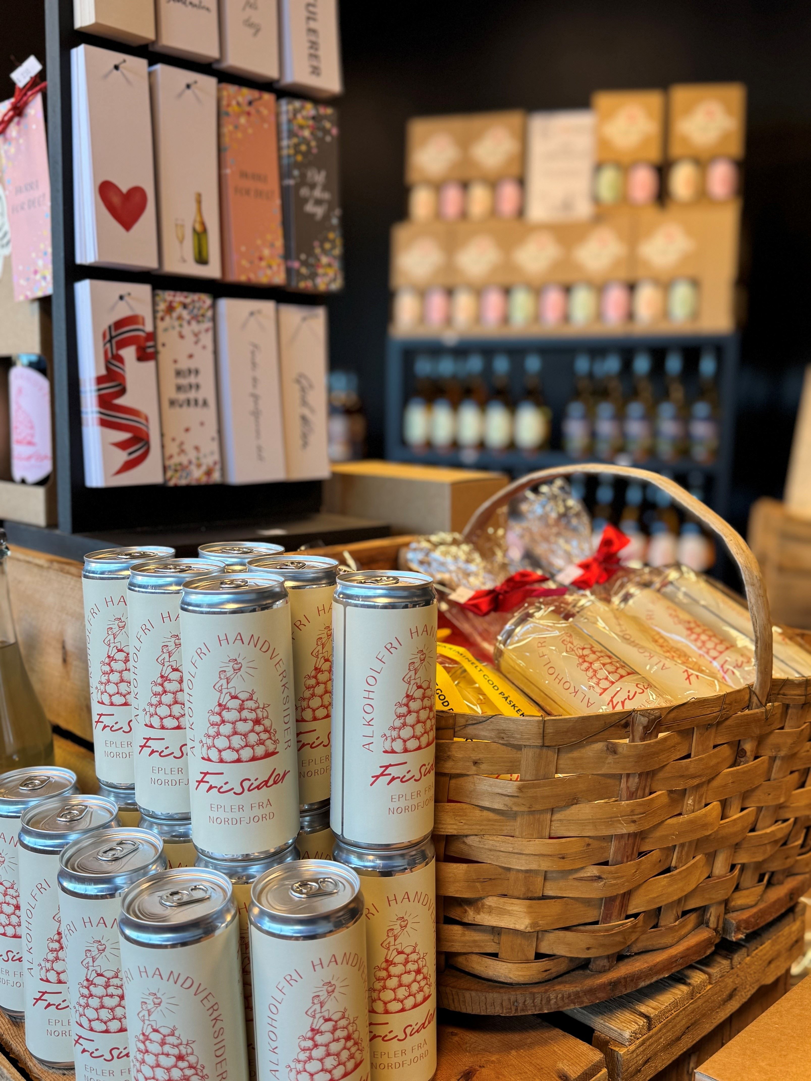 Cans of craft cider stacked beside a wicker gift basket with wrapped items, with shelves of cards and bottles behind.