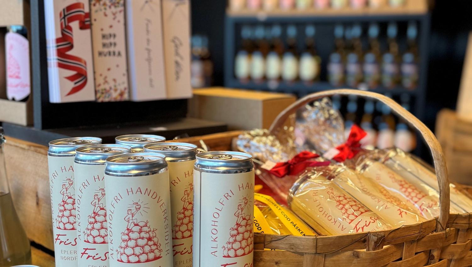 Cans of craft cider stacked beside a wicker gift basket with wrapped items, with shelves of cards and bottles behind.