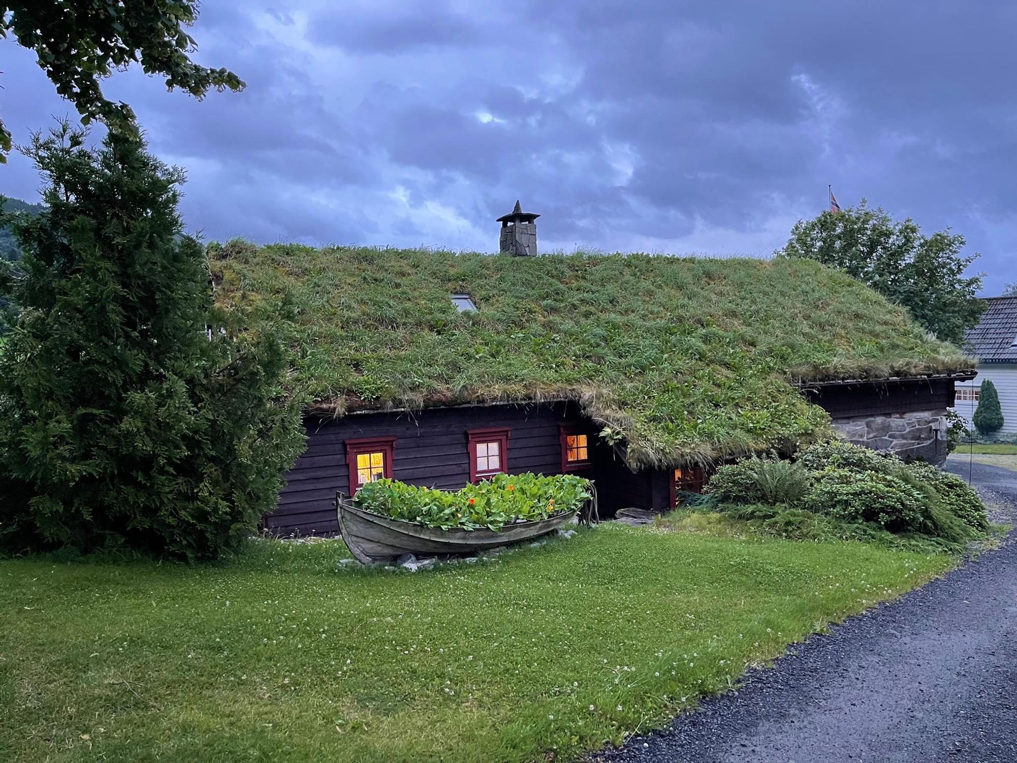 Orheimstunet in Nordfjord - A dark wooden cabin with a grass-covered roof and lit windows, with a rowboat planter on a lawn under cloudy sky..