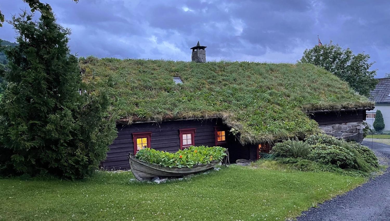 Orheimstunet in Nordfjord - A dark wooden cabin with a grass-covered roof and lit windows, with a rowboat planter on a lawn under cloudy sky..