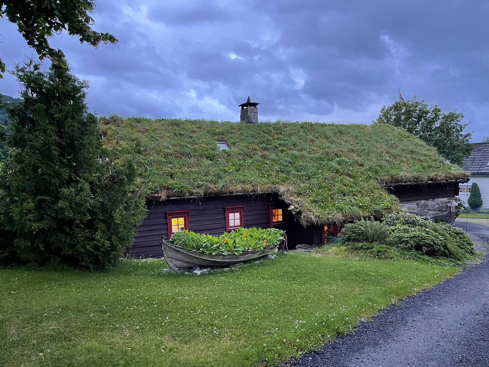 Orheimstunet in Nordfjord - A dark wooden cabin with a grass-covered roof and lit windows, with a rowboat planter on a lawn under cloudy sky..