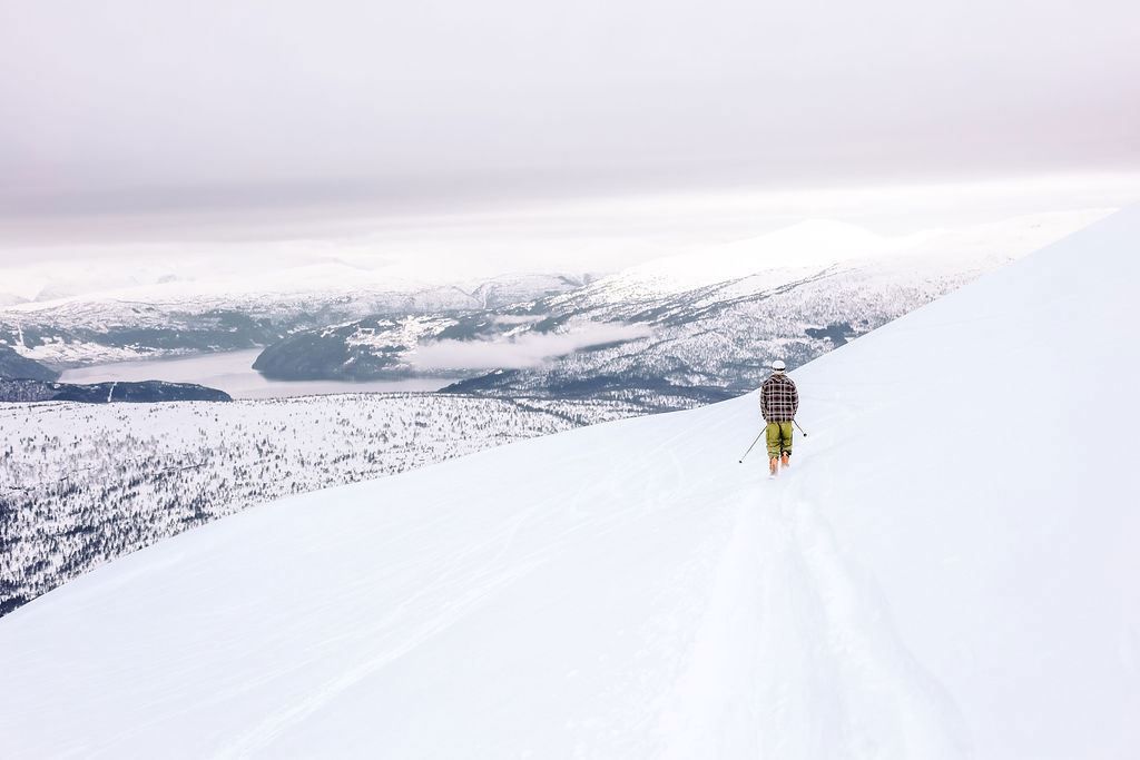 A lone skier with poles walks along a snowy ridgeline above distant mountains and a lake under an overcast sky.