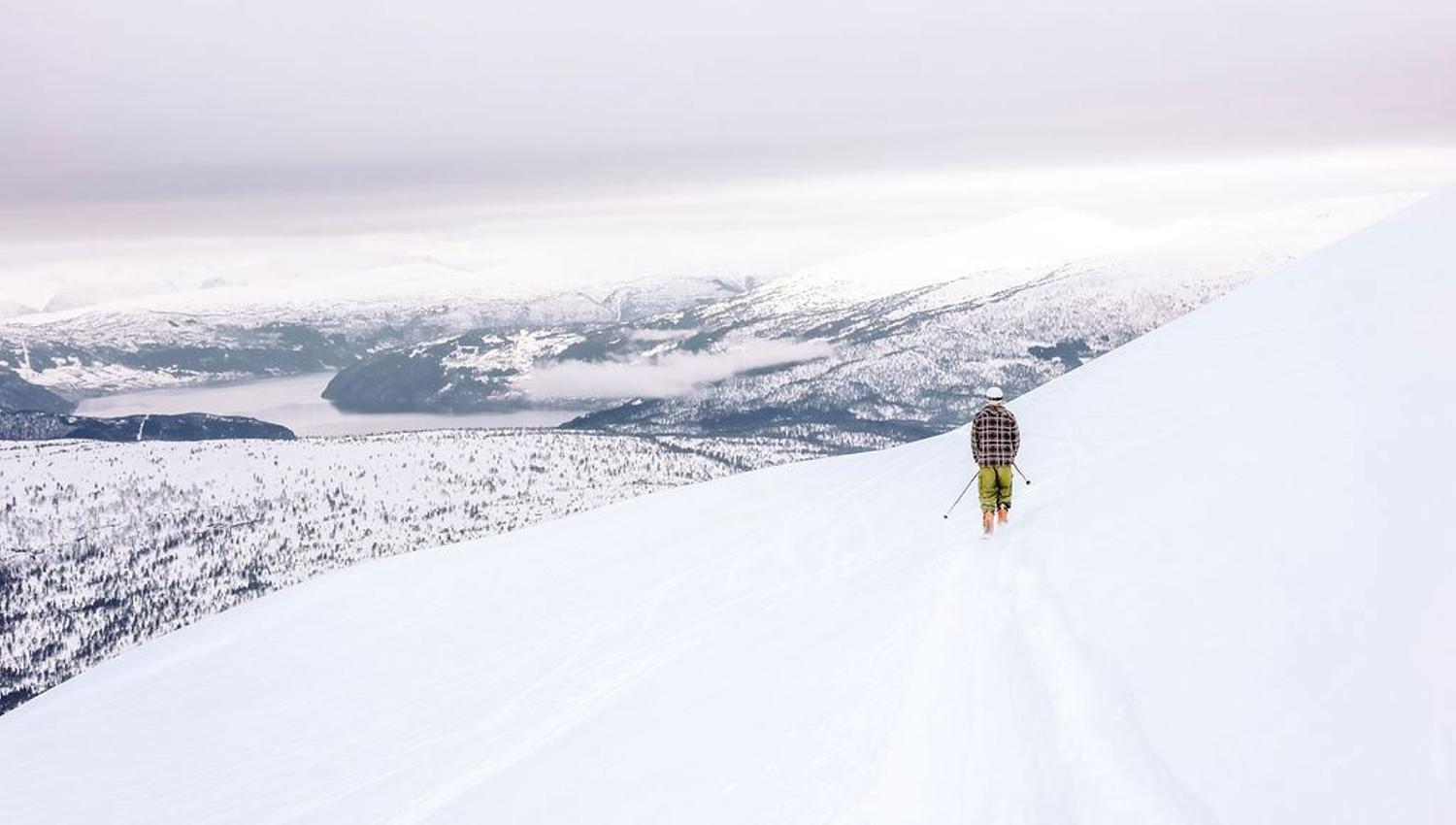 A lone skier with poles walks along a snowy ridgeline above distant mountains and a lake under an overcast sky.