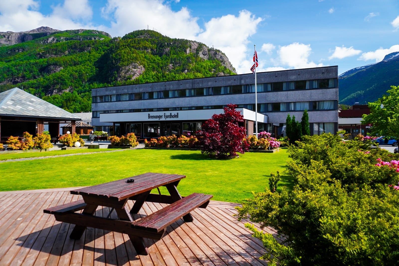 Grått hotellbygg ved grønt plenområde med piknikbord, buskar og norsk flagg, med fjell og blå himmel bak..