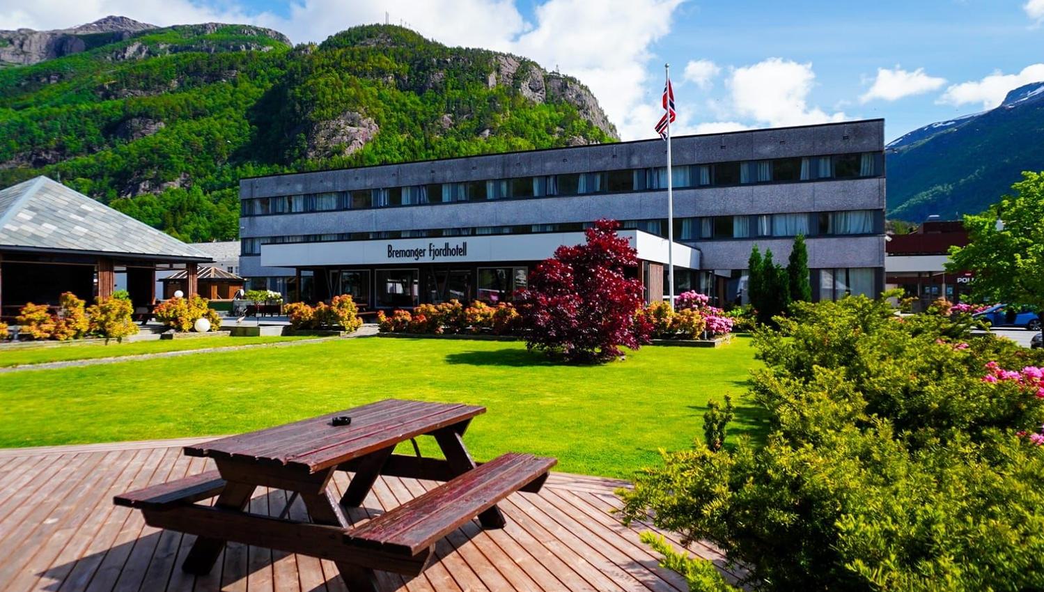 Grått hotellbygg ved grønt plenområde med piknikbord, buskar og norsk flagg, med fjell og blå himmel bak..