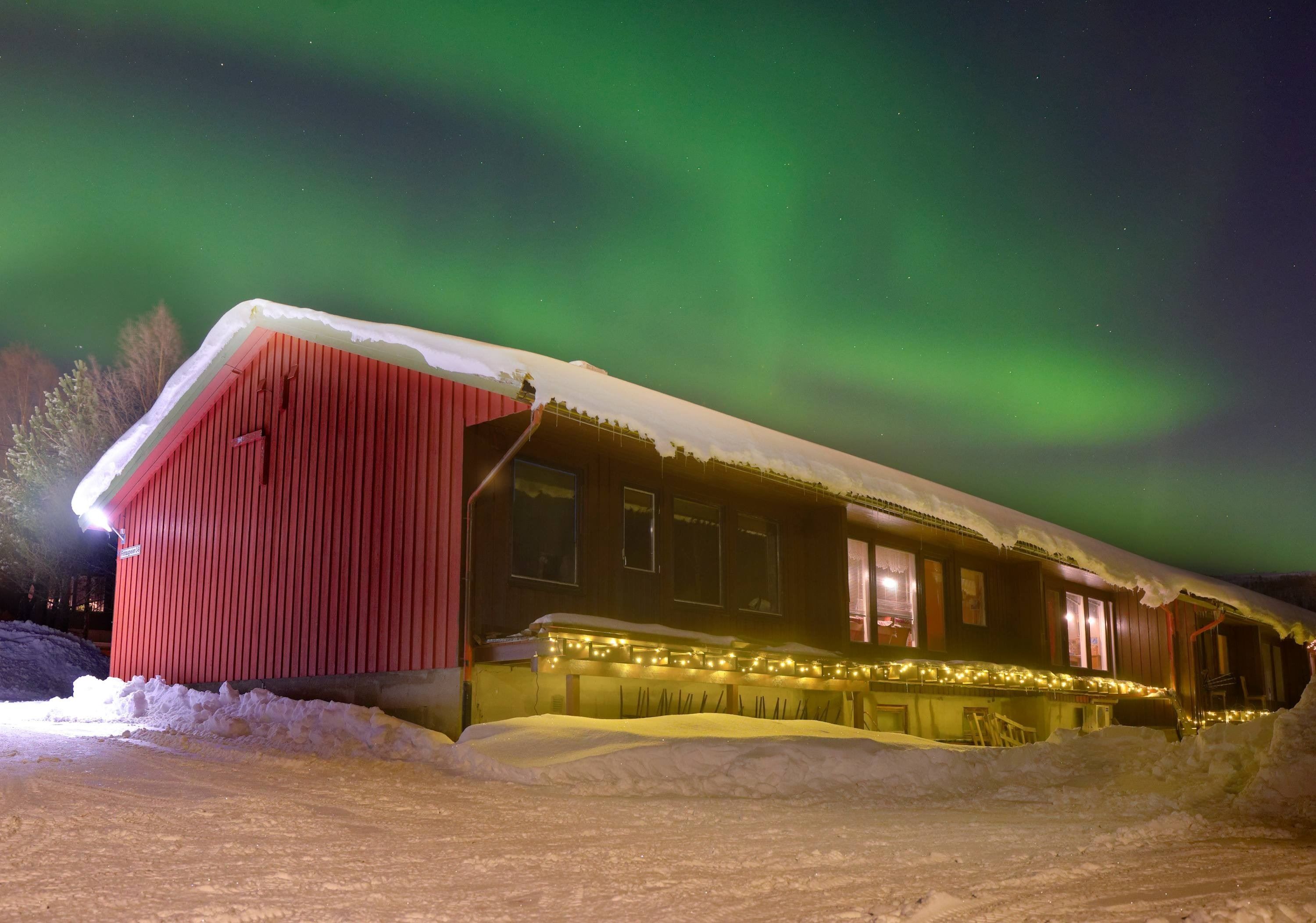 Red wooden cabin with lit windows and string lights in deep snow, beneath green aurora in a dark sky.