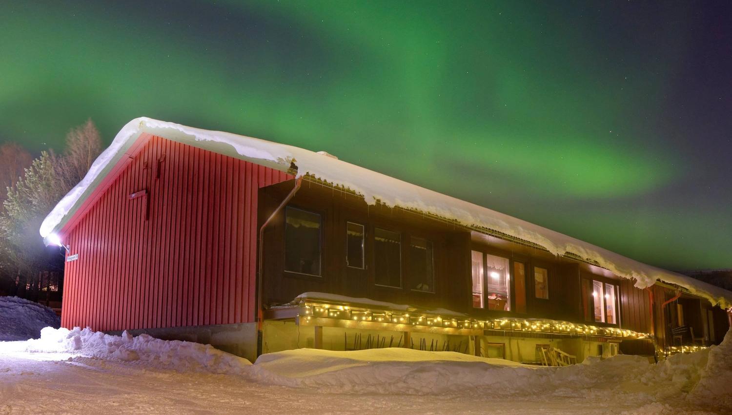 Red wooden cabin with lit windows and string lights in deep snow, beneath green aurora in a dark sky.