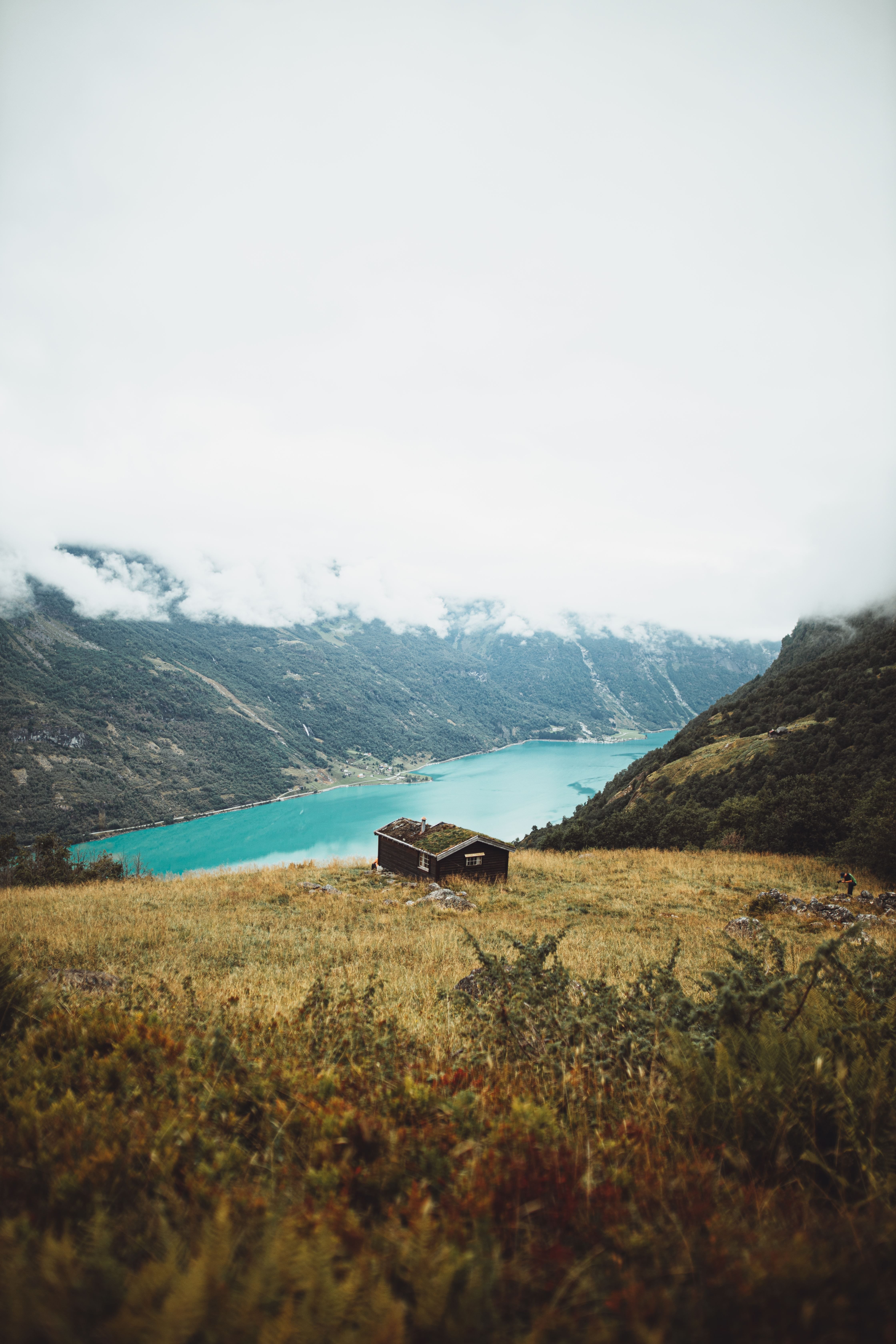 Small wooden cabin on a grassy hillside on Kvamssetra overlooking a turquoise fjord and misty mountains under low clouds..