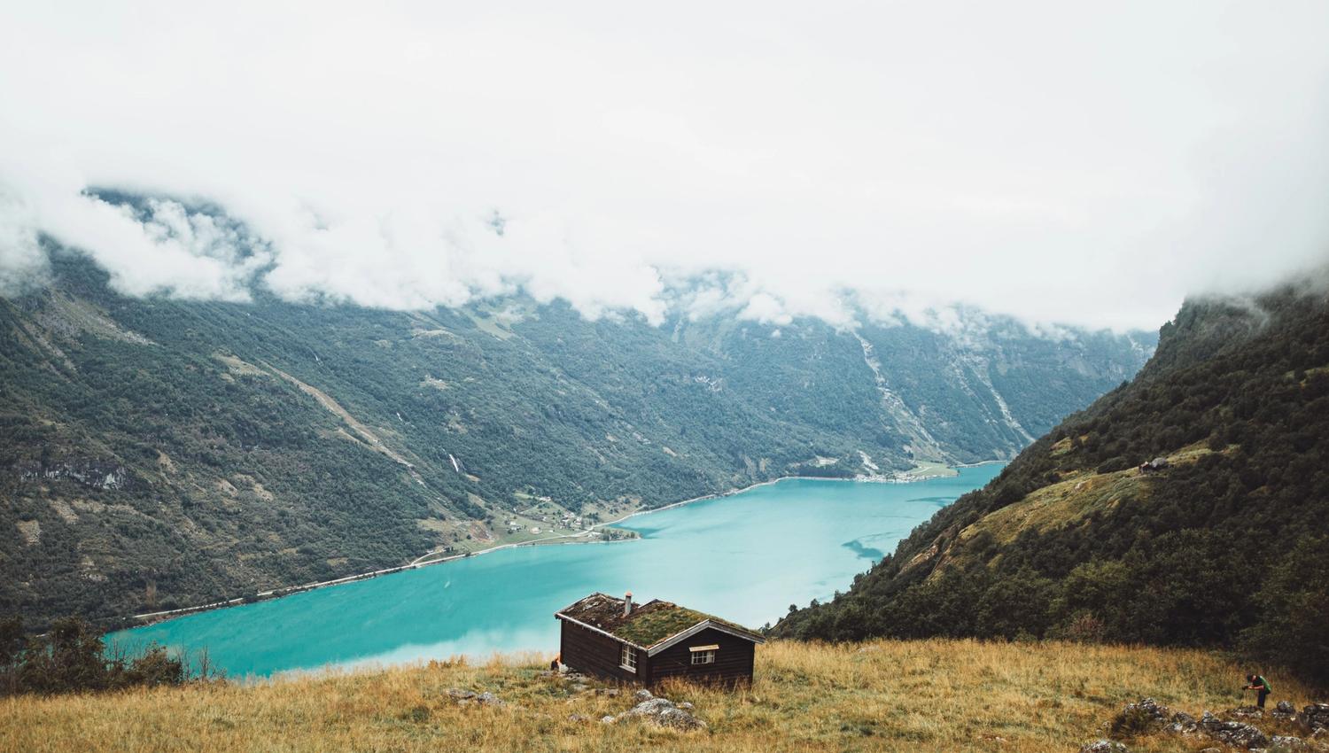 Small wooden cabin on a grassy hillside on Kvamssetra overlooking a turquoise fjord and misty mountains under low clouds..