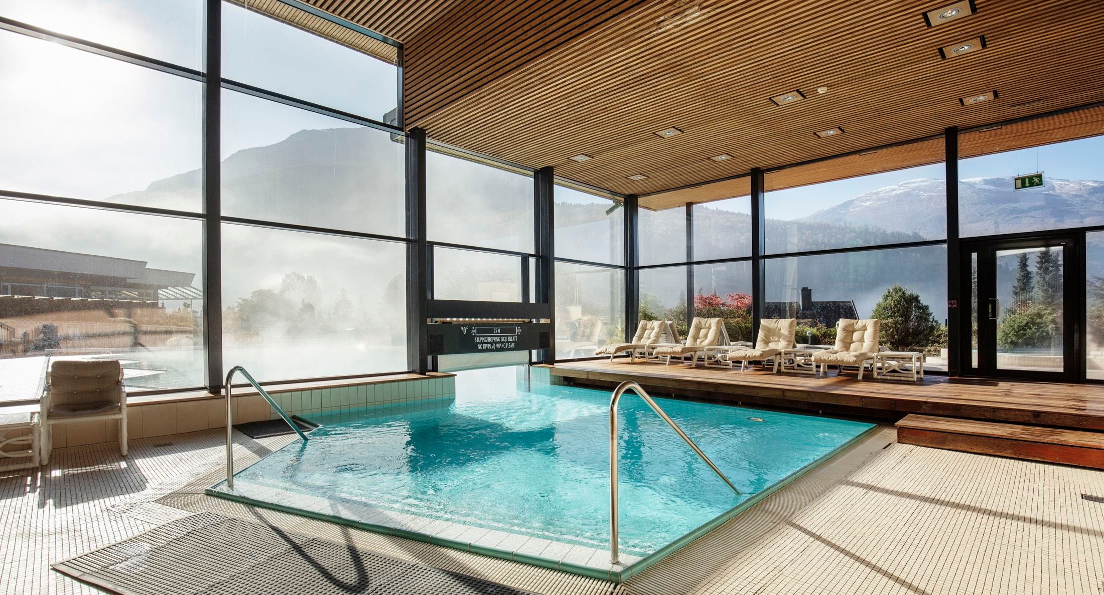 Sunlit indoor pool with metal handrails, lounge chairs on a wooden deck, and large windows facing misty mountains at Hotel Alexandra..