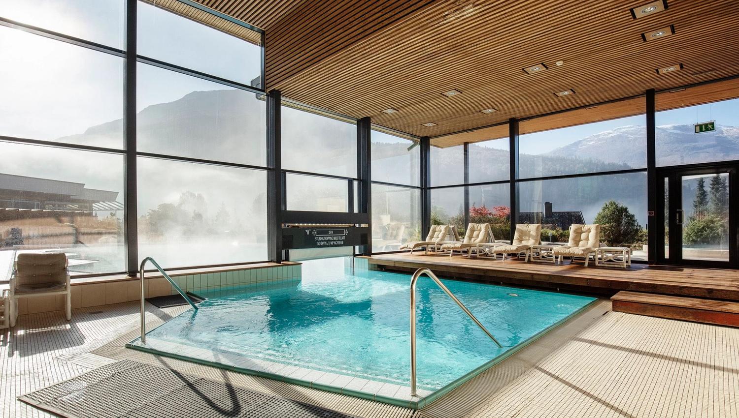 Sunlit indoor pool with metal handrails, lounge chairs on a wooden deck, and large windows facing misty mountains at Hotel Alexandra..