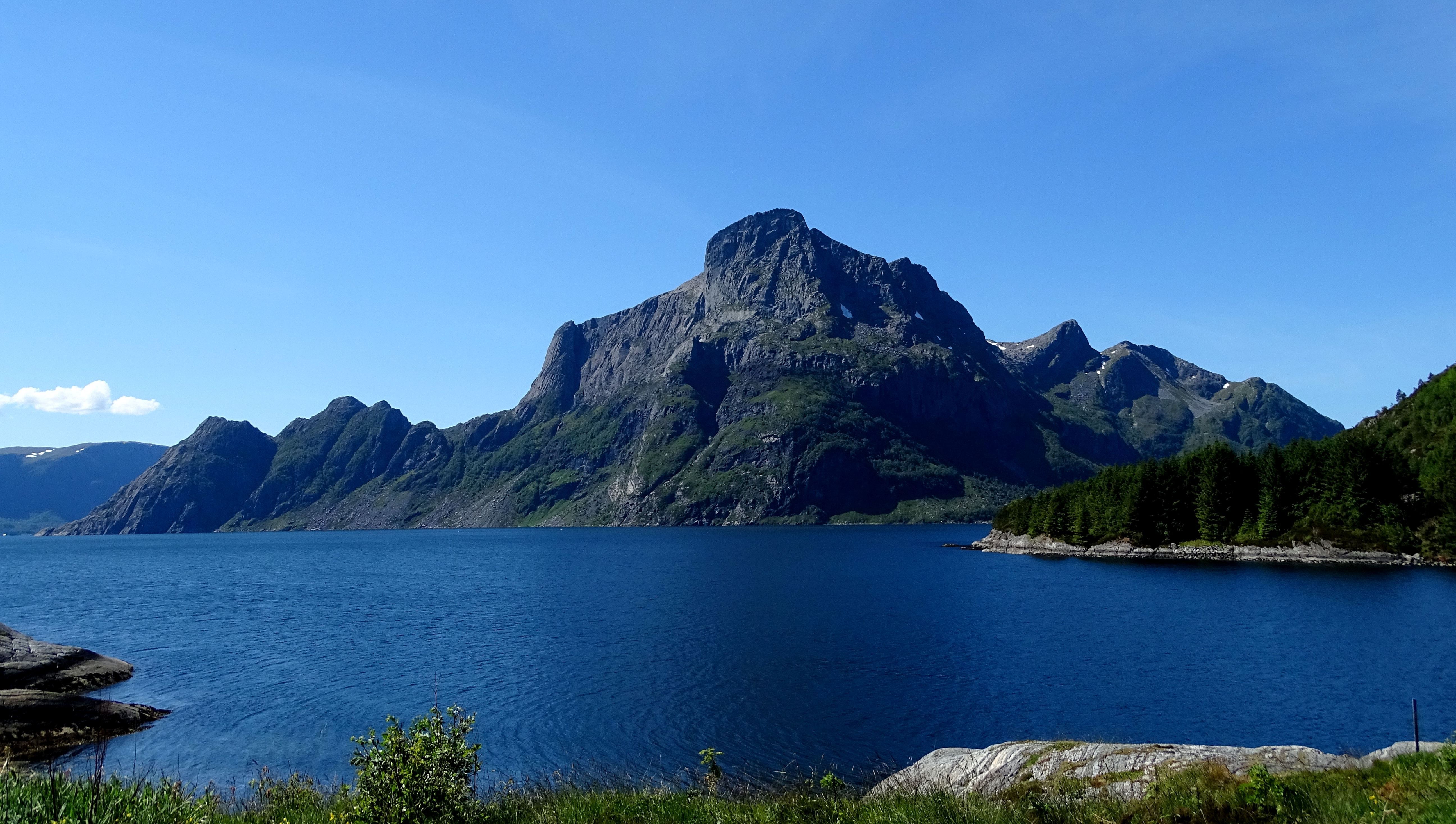 Opplevingar i nærleiken av Bremanger Fjordhotell: Stort fjellmassiv Hornelen reiser seg bak ei blå fjord, med grøne skråningar og skogkledd nes under klår himmel..