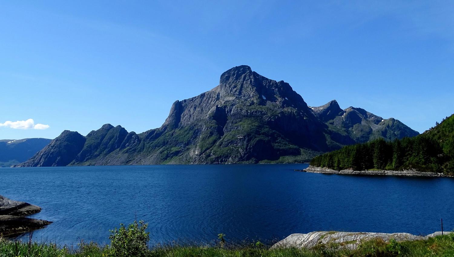 Opplevingar i nærleiken av Bremanger Fjordhotell: Stort fjellmassiv Hornelen reiser seg bak ei blå fjord, med grøne skråningar og skogkledd nes under klår himmel..
