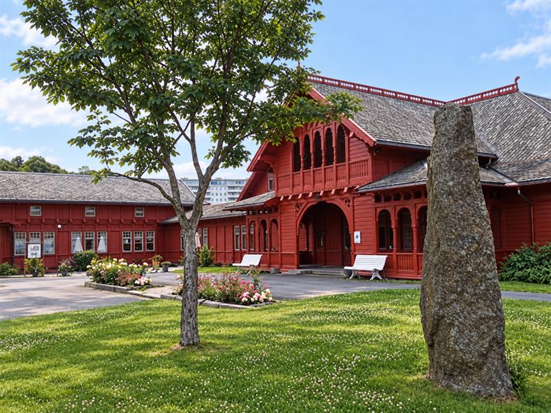 Historic red wooden spa building in Sandefjord viewed from the lawn, with a leafy tree, a tall standing stone and flower beds.