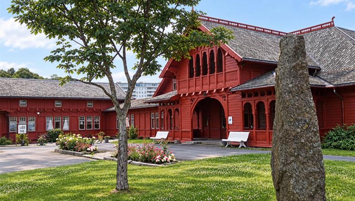 Historic red wooden spa building in Sandefjord viewed from the lawn, with a leafy tree, a tall standing stone and flower beds.