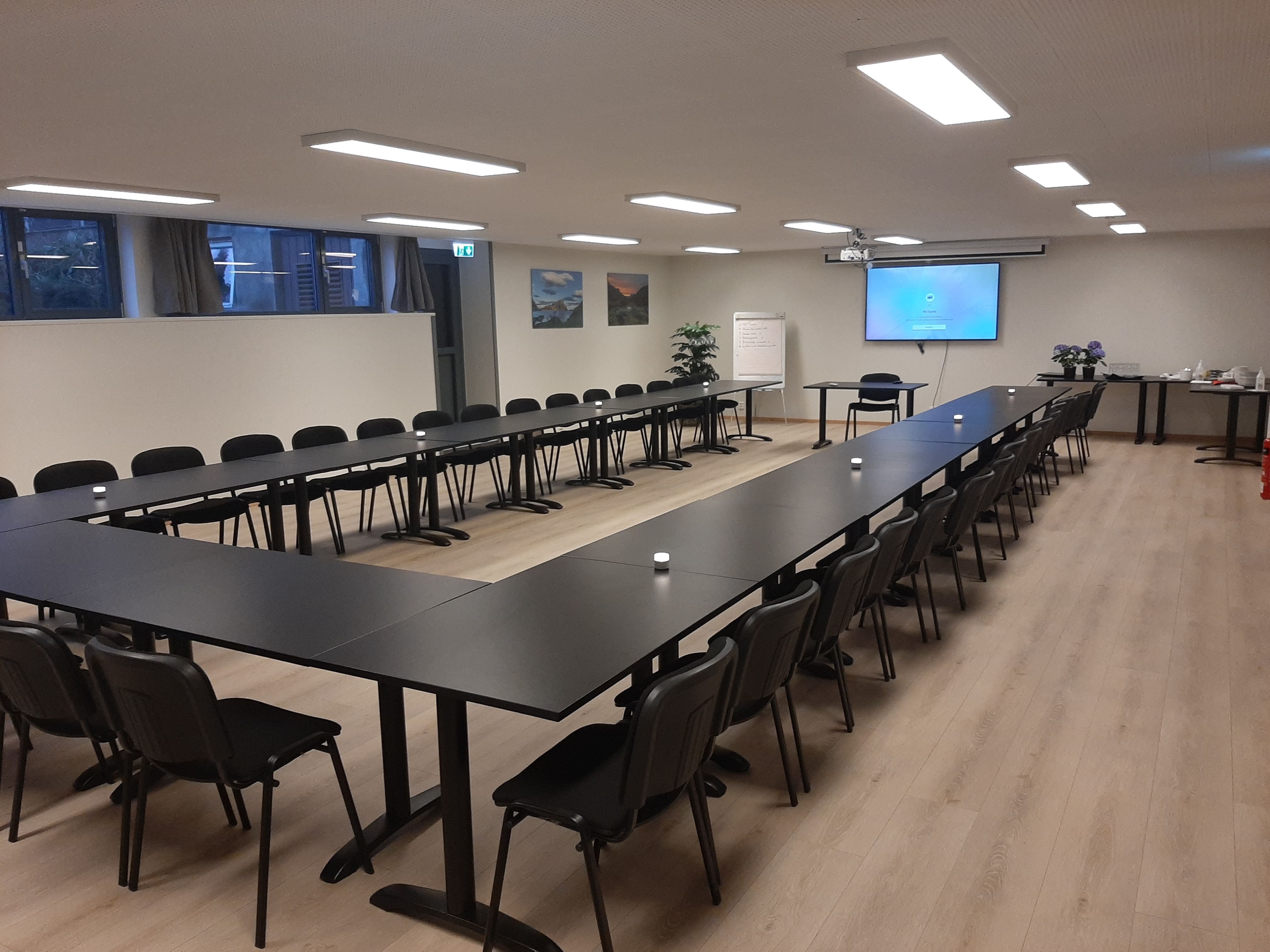 Stort møterom med U-forma svarte bord og stolar, skjerm på veggen, flippover og serveringsbord langs sida..